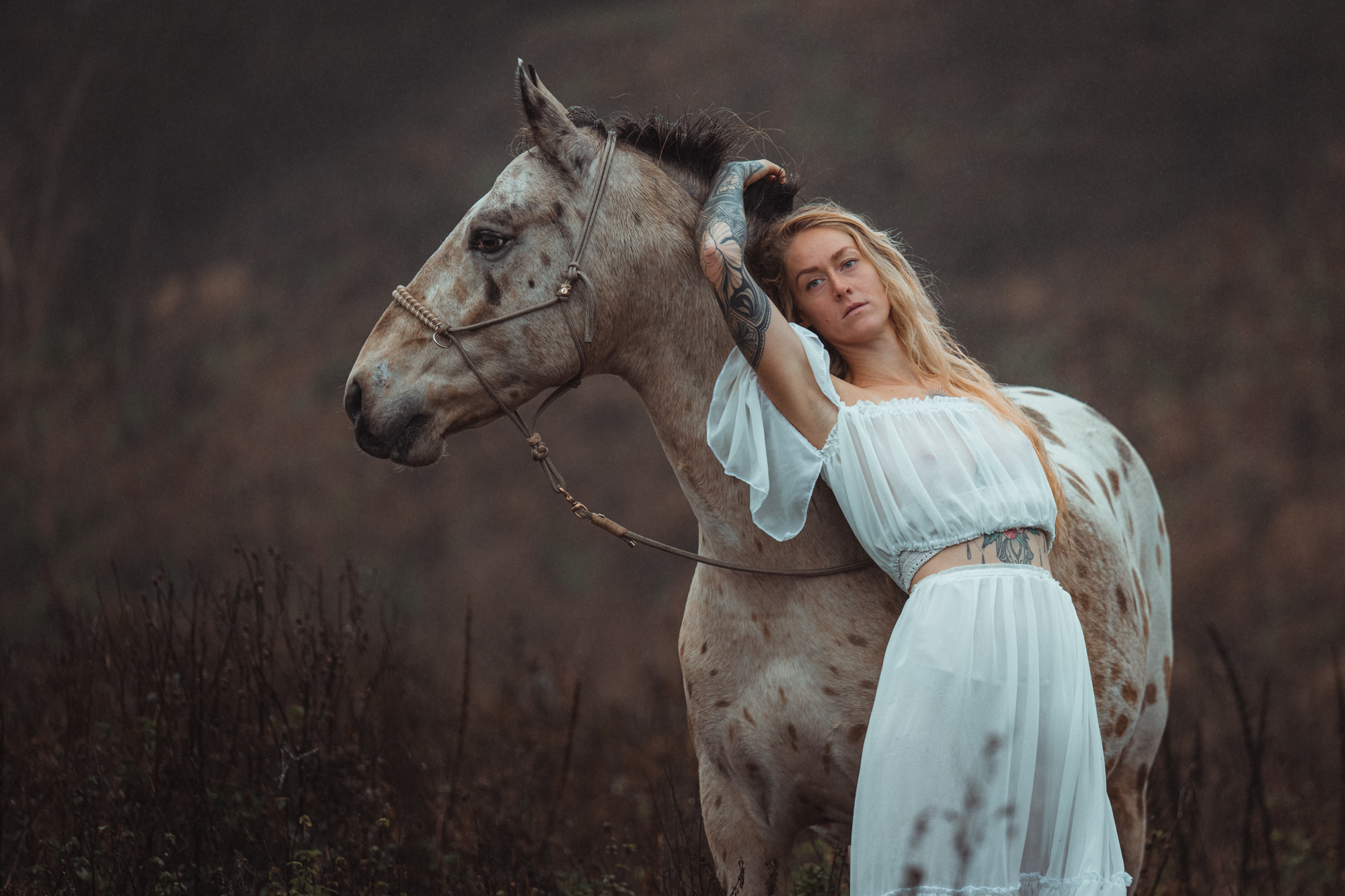 Frau in weißem Kleid lehnt an einem scheckigen Pferd in nebliger Landschaft